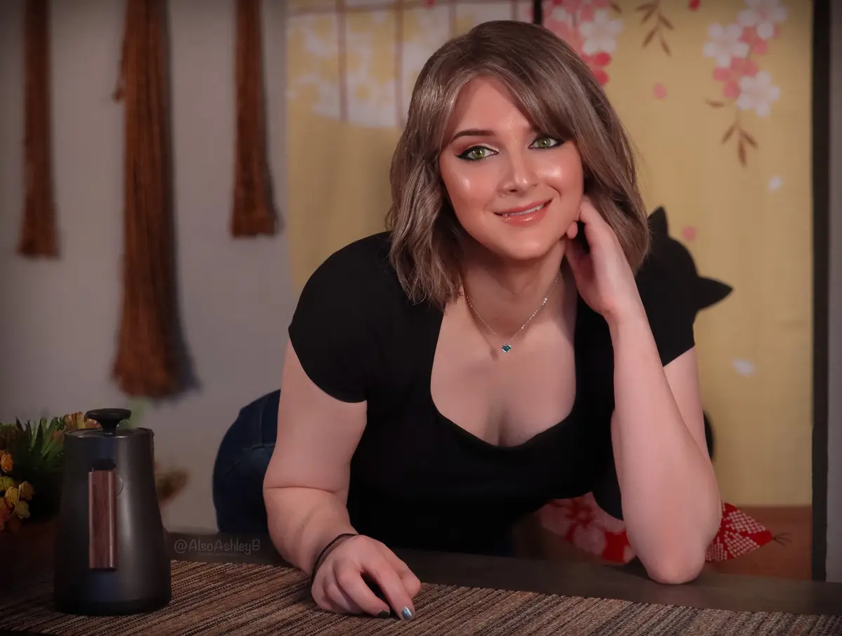A woman with short brown hair and green eyes smiles while leaning on a table, wearing a black top and a heart-shaped…