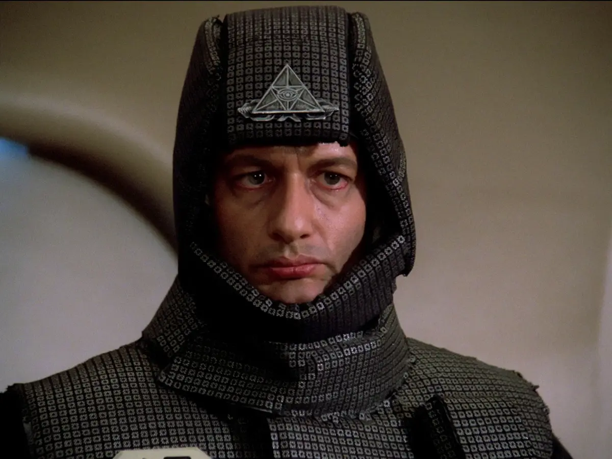 A man in a dark, patterned hooded outfit with a triangular eye emblem on his forehead stares intensely at the camera. The…