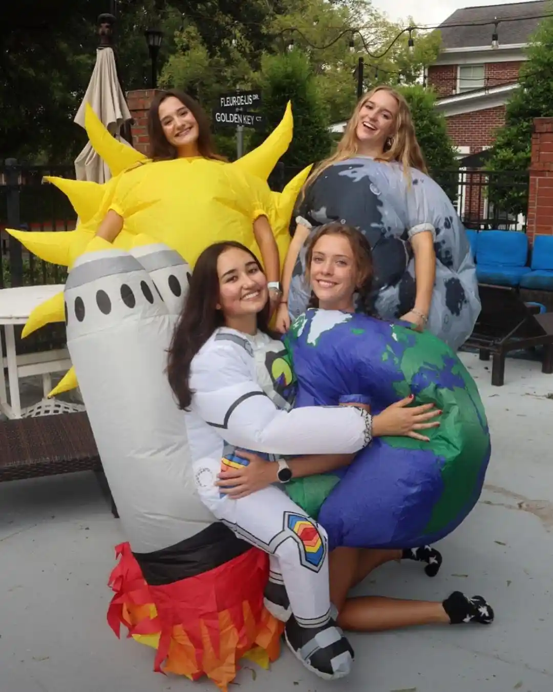 Four young women pose outdoors in inflatable costumes: one as the sun with yellow spikes, one as the moon with gray and…