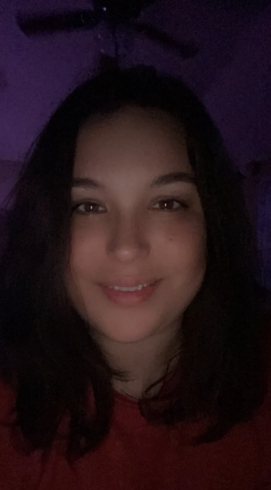 A dimly lit selfie of a woman with dark hair and a red shirt, smiling slightly while looking at the camera. The background…