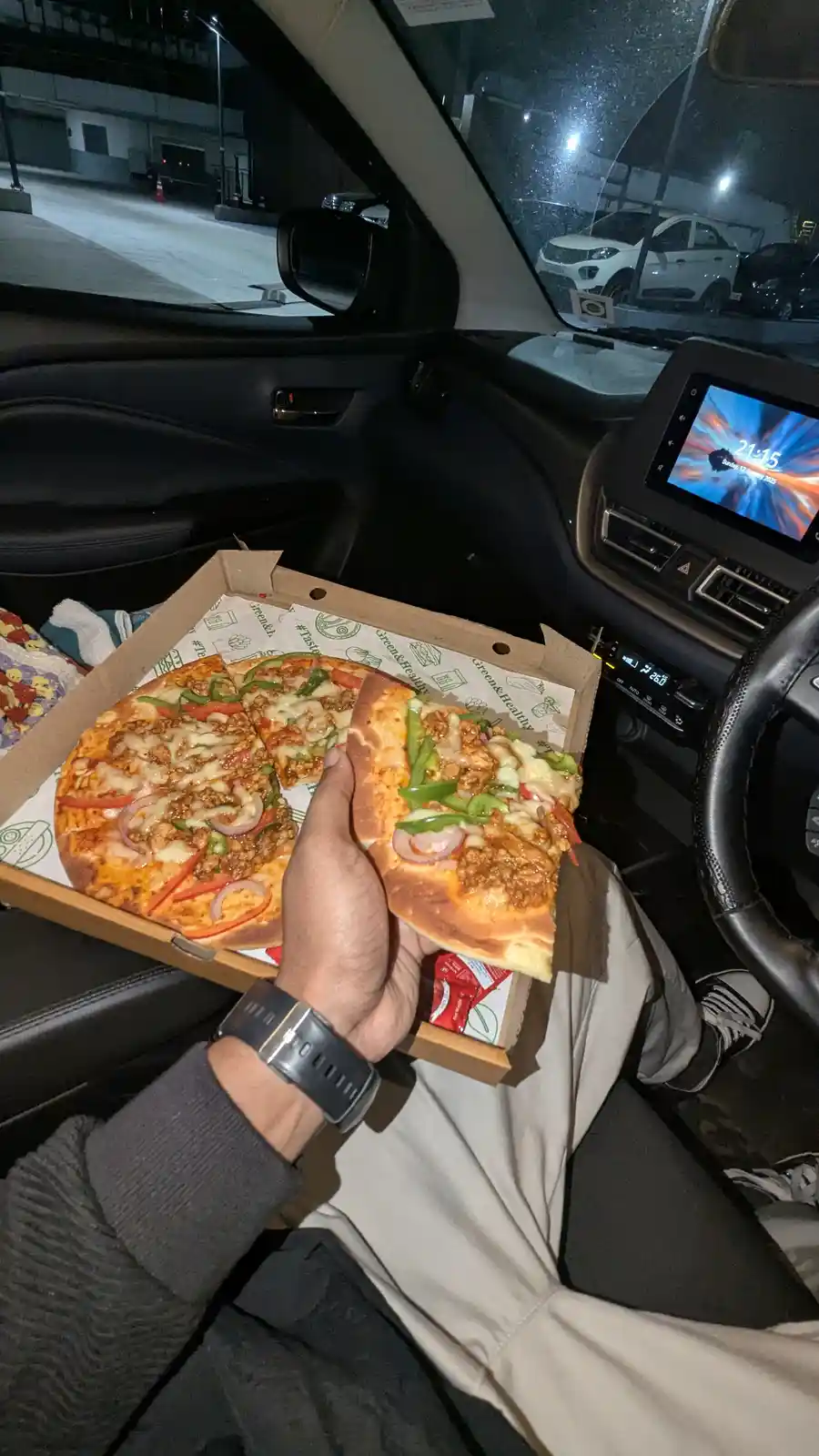 A person sits in the driver's seat of a car at night, holding a slice of pizza from an open cardboard box. The car's…