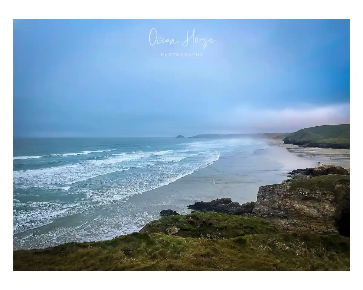 A scenic coastal photograph showing a wide beach with waves rolling in under a hazy blue sky, viewed from a grassy cliff…