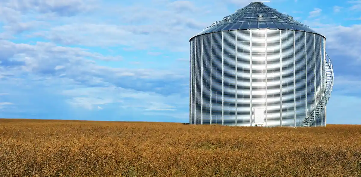 A large, silver, cylindrical metal grain silo stands alone in a vast field of golden, harvested crops under a partly cloudy…