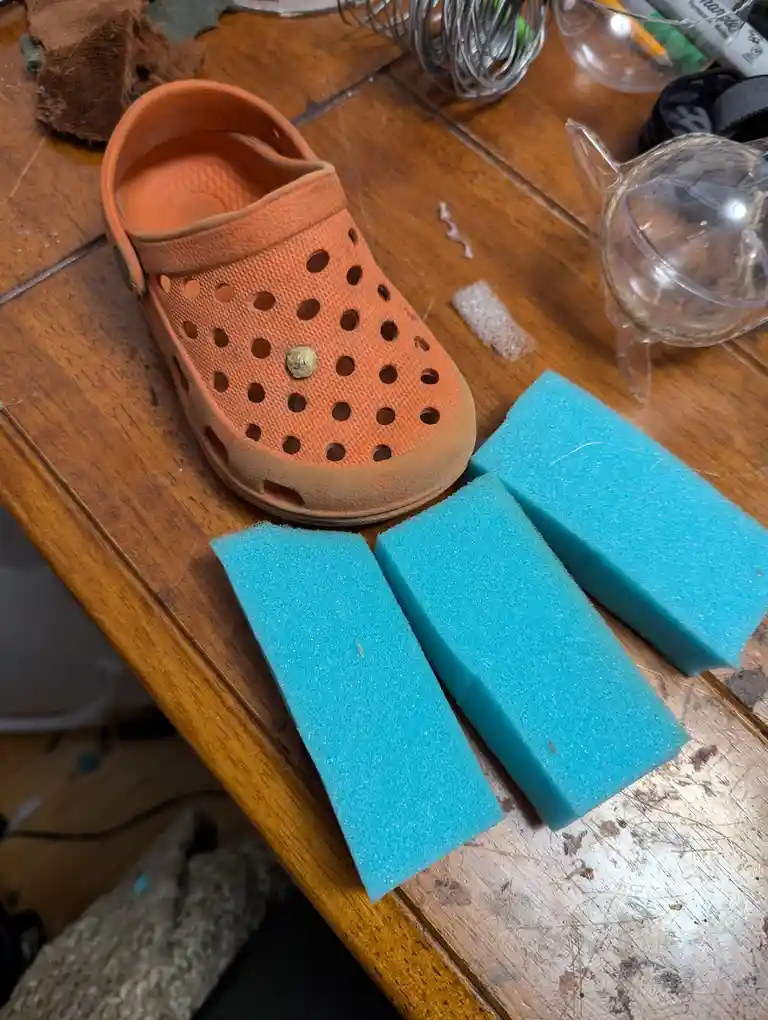 An orange Croc clog sits on a wooden table next to three rectangular blue sponges, with various craft supplies in the…