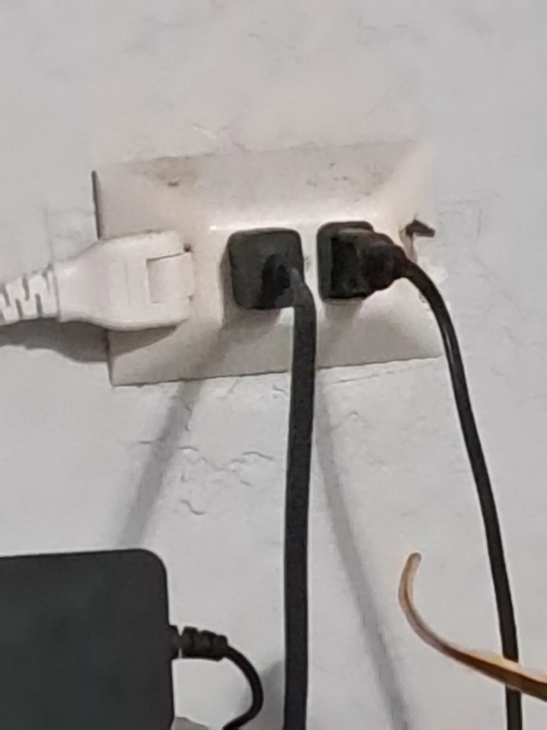 A close-up photo shows a white electrical outlet with three plugs inserted: a white USB-style plug on the left, and two…