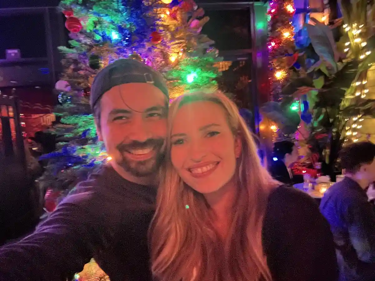A selfie of a smiling man and woman posing in front of a brightly lit, multi-colored Christmas tree in a festive, dimly lit…