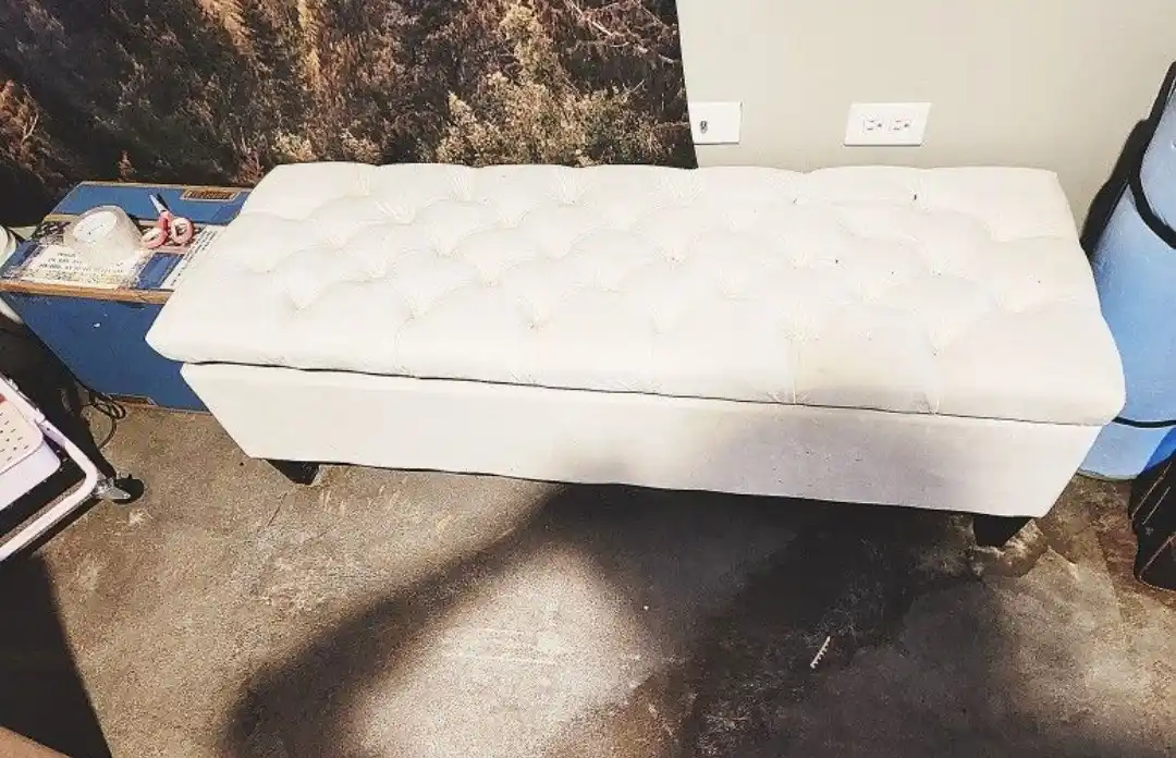 A white tufted ottoman sits on a concrete floor in a cluttered room, surrounded by various items including a blue cart with…