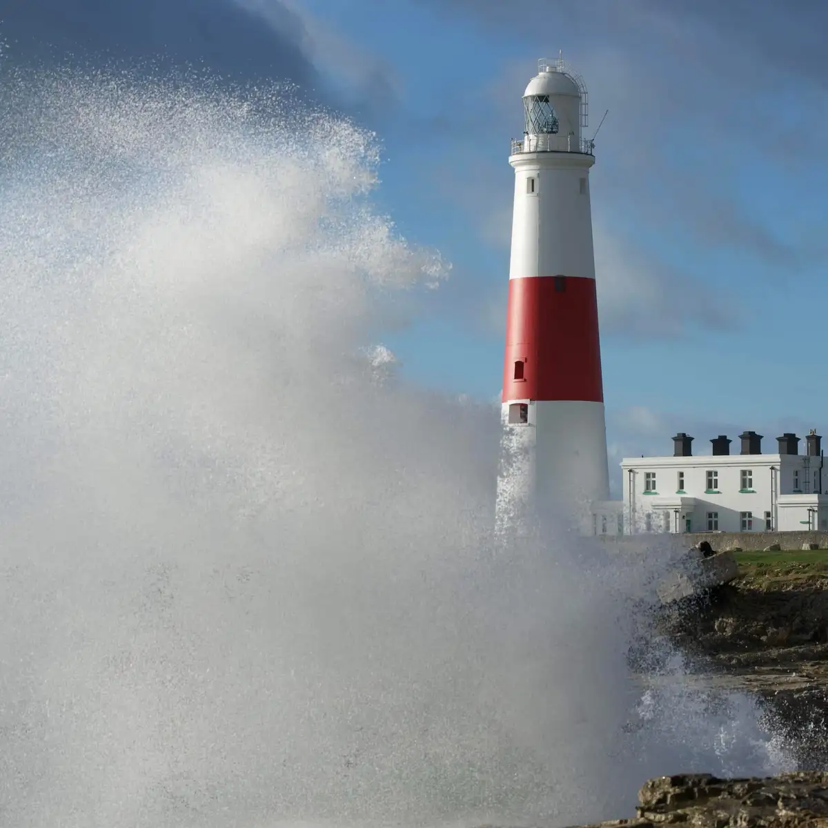 A tall white lighthouse with a red band is shown standing firm as a massive wave crashes against its base, sending a huge…