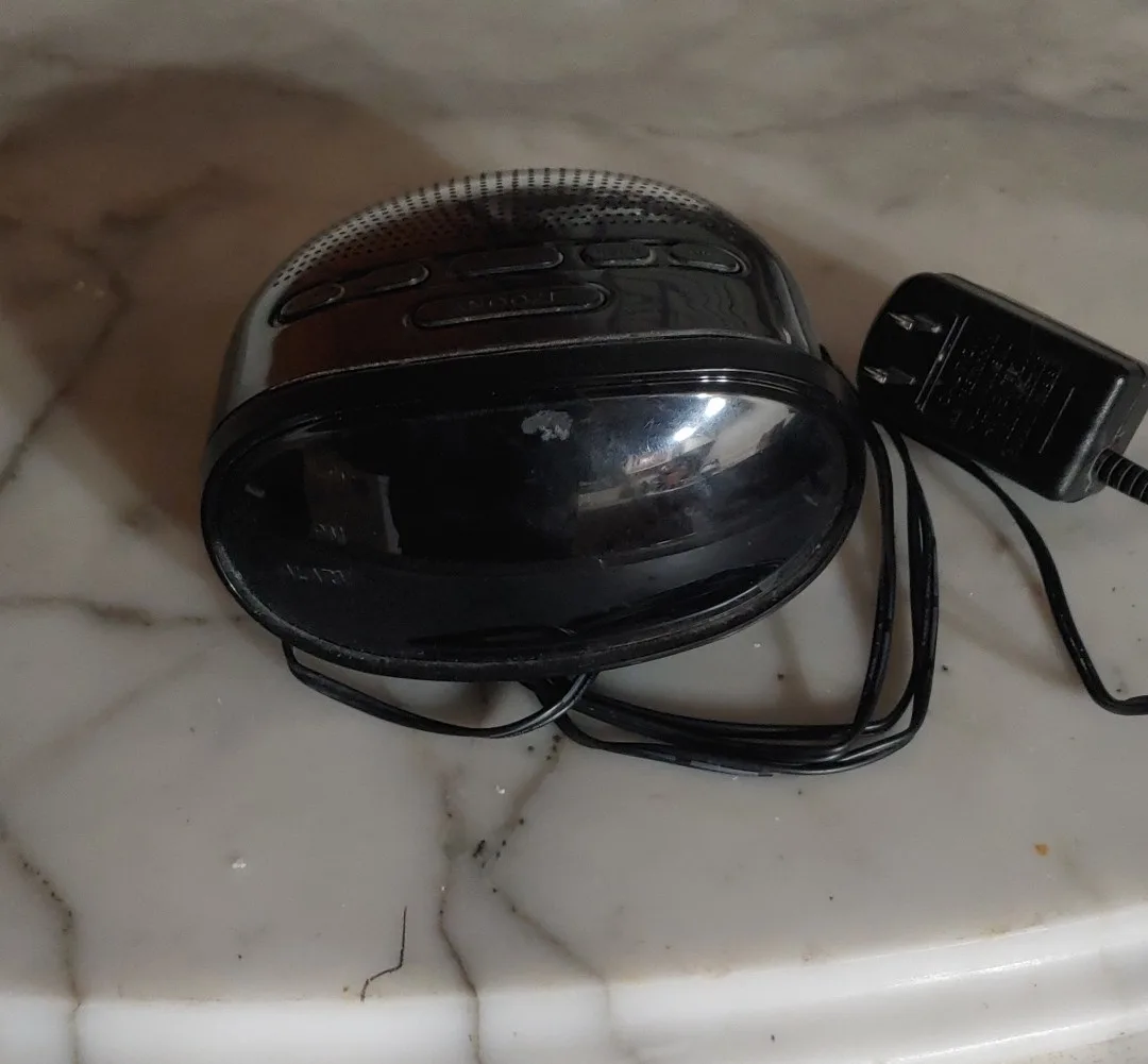 A black, oval-shaped, glossy electronic device with buttons and a power cord lies on a white marble surface. The overlaid…
