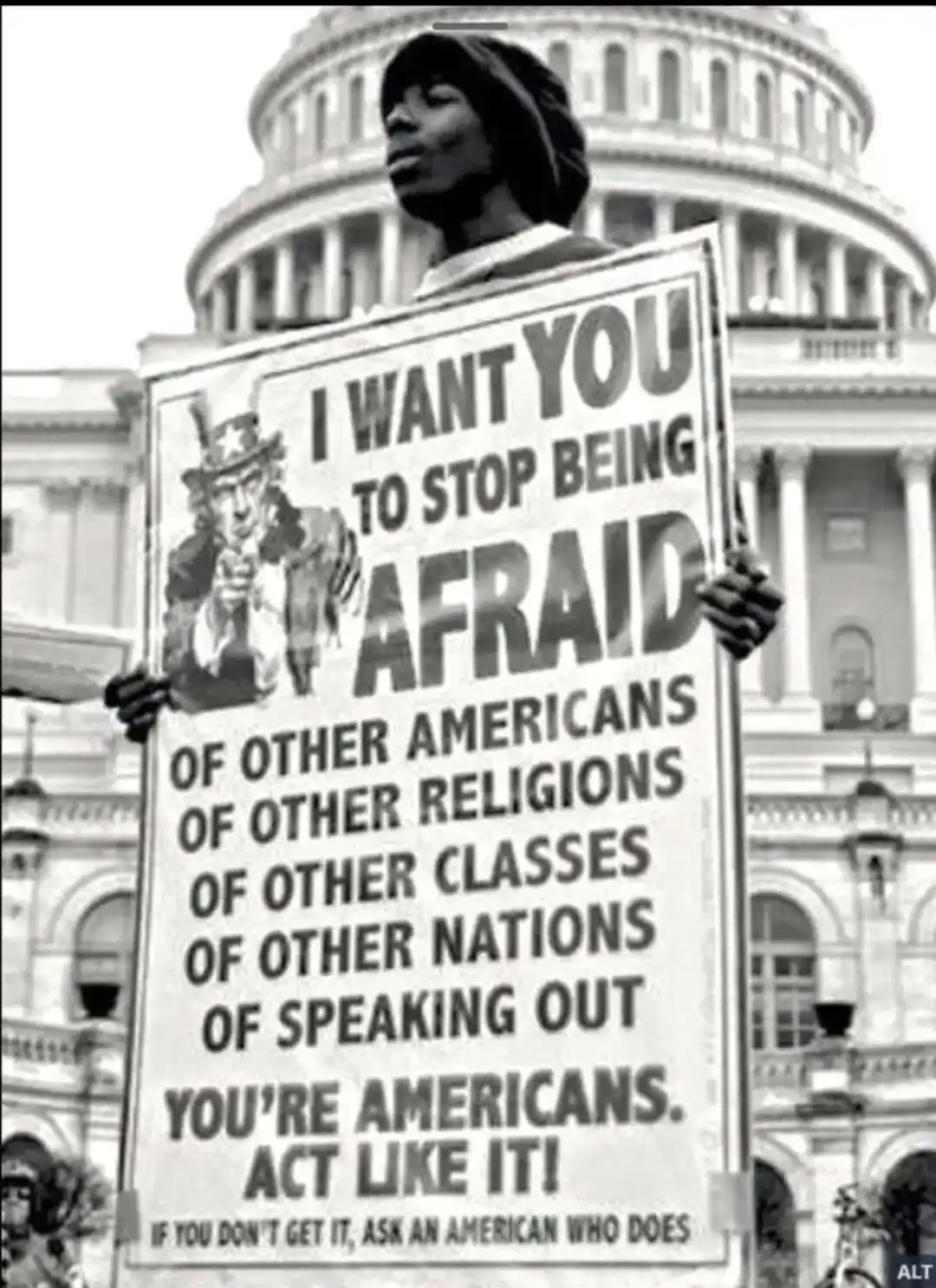 A black-and-white photo shows a person holding a large protest sign in front of the U.S. Capitol building. The sign features…