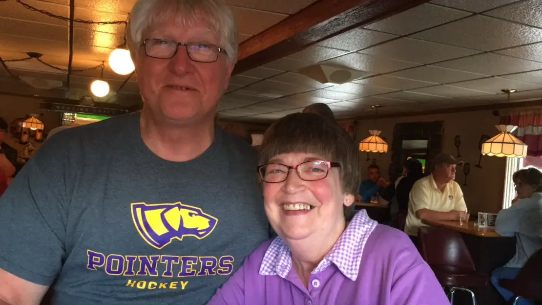 An older man and woman pose for a photo in a restaurant. The man wears a gray t-shirt with a yellow and purple logo of a…