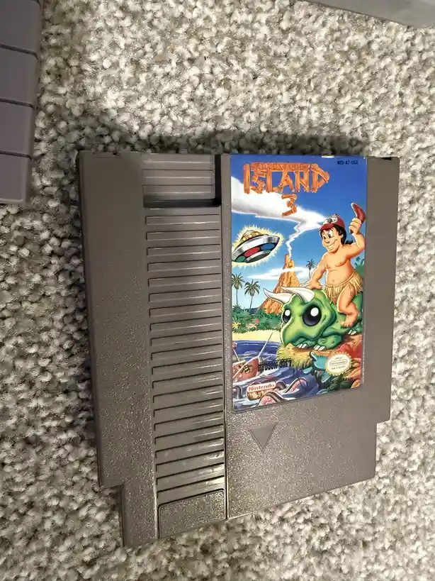 A close-up photo of a gray Nintendo Entertainment System game cartridge lying on a textured gray carpet. The label shows the…