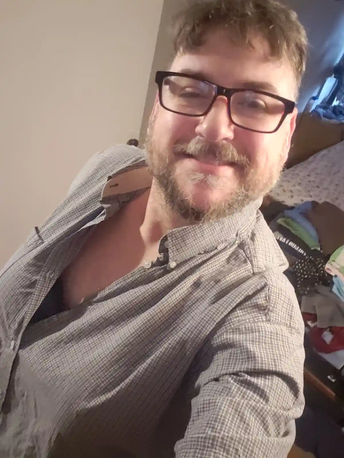 A man with glasses and a beard smiles at the camera while wearing a plaid shirt with the top buttons undone, revealing his…