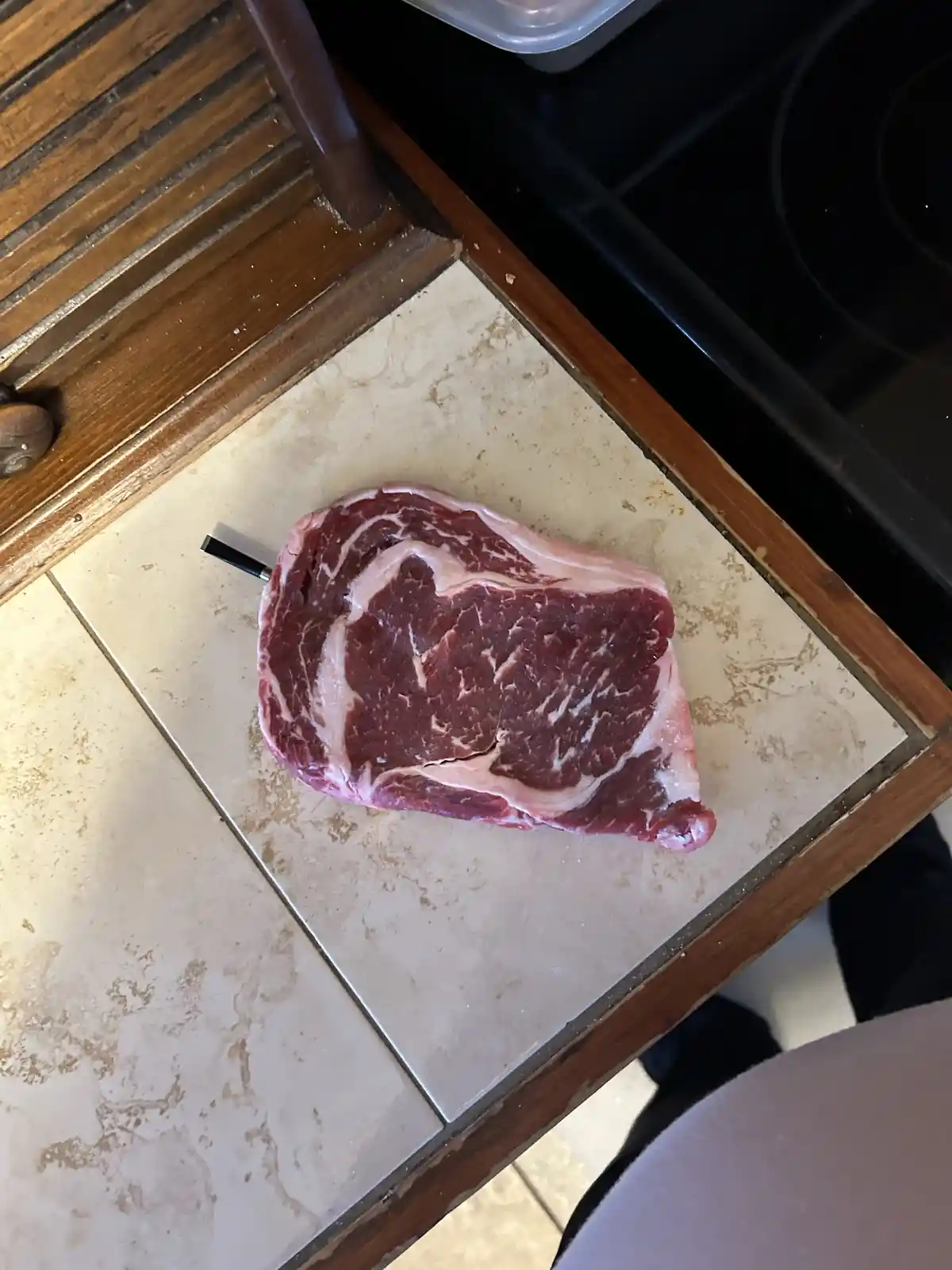 A raw, marbled steak sits on a beige tiled kitchen counter with a black-handled knife stuck into it. The background shows a…