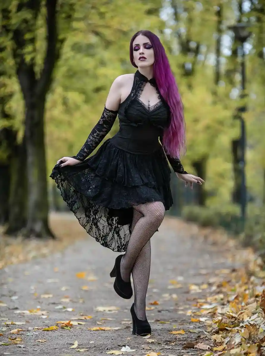 A woman with long purple hair and dramatic makeup poses in a black lace dress, fishnet stockings, and platform heels on a…