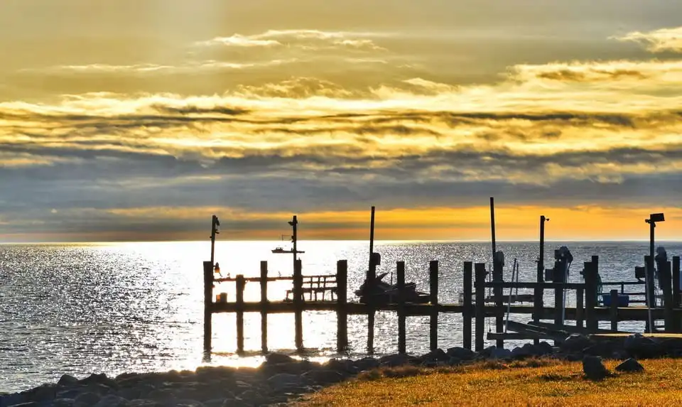A serene sunset over water with a weathered wooden pier silhouetted against golden clouds and shimmering waves. The overlaid…