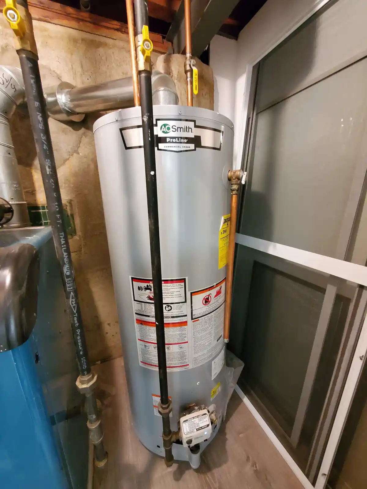 A gray AO Smith ProLine commercial-grade water heater is shown in a basement, surrounded by copper and black pipes, with a…