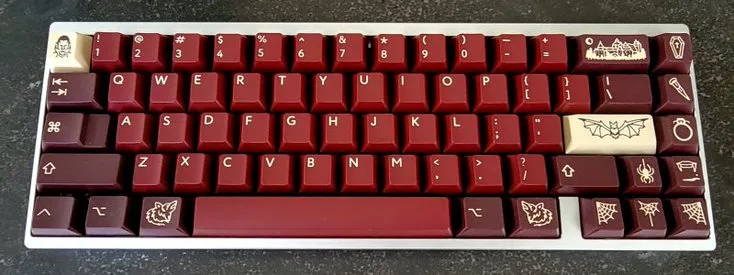 A mechanical keyboard with red and burgundy keycaps, many of which are customized with white engravings of gothic and…