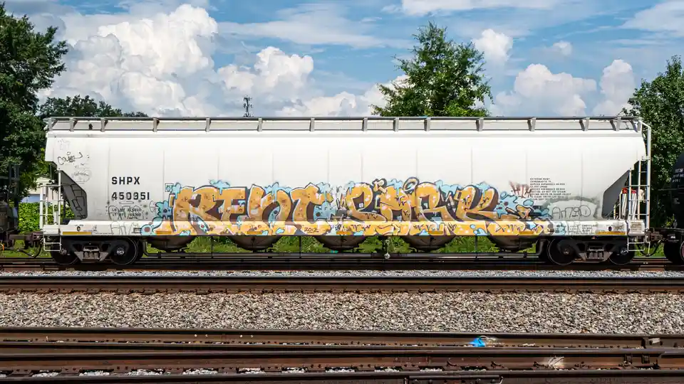 A white freight train car with large yellow and blue graffiti reading 'RELOADED' sits on railroad tracks under a partly…