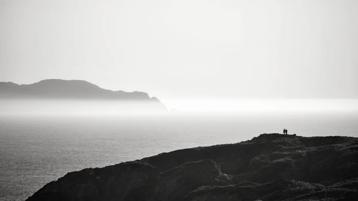 A black-and-white photo shows two small human figures standing on a grassy cliff overlooking a vast, fog-covered ocean with…
