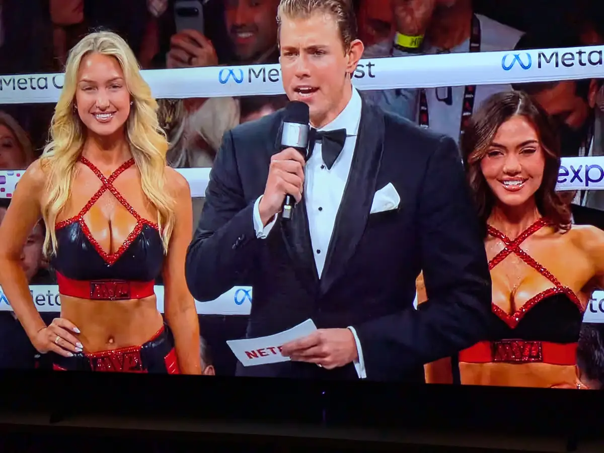 A man in a tuxedo holding a microphone and a piece of paper with 'NETFLIX' written on it stands between two women in red and…