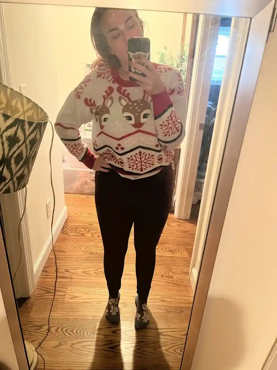 A person takes a mirror selfie wearing a festive white sweater with red reindeer and snowflake patterns, paired with black…