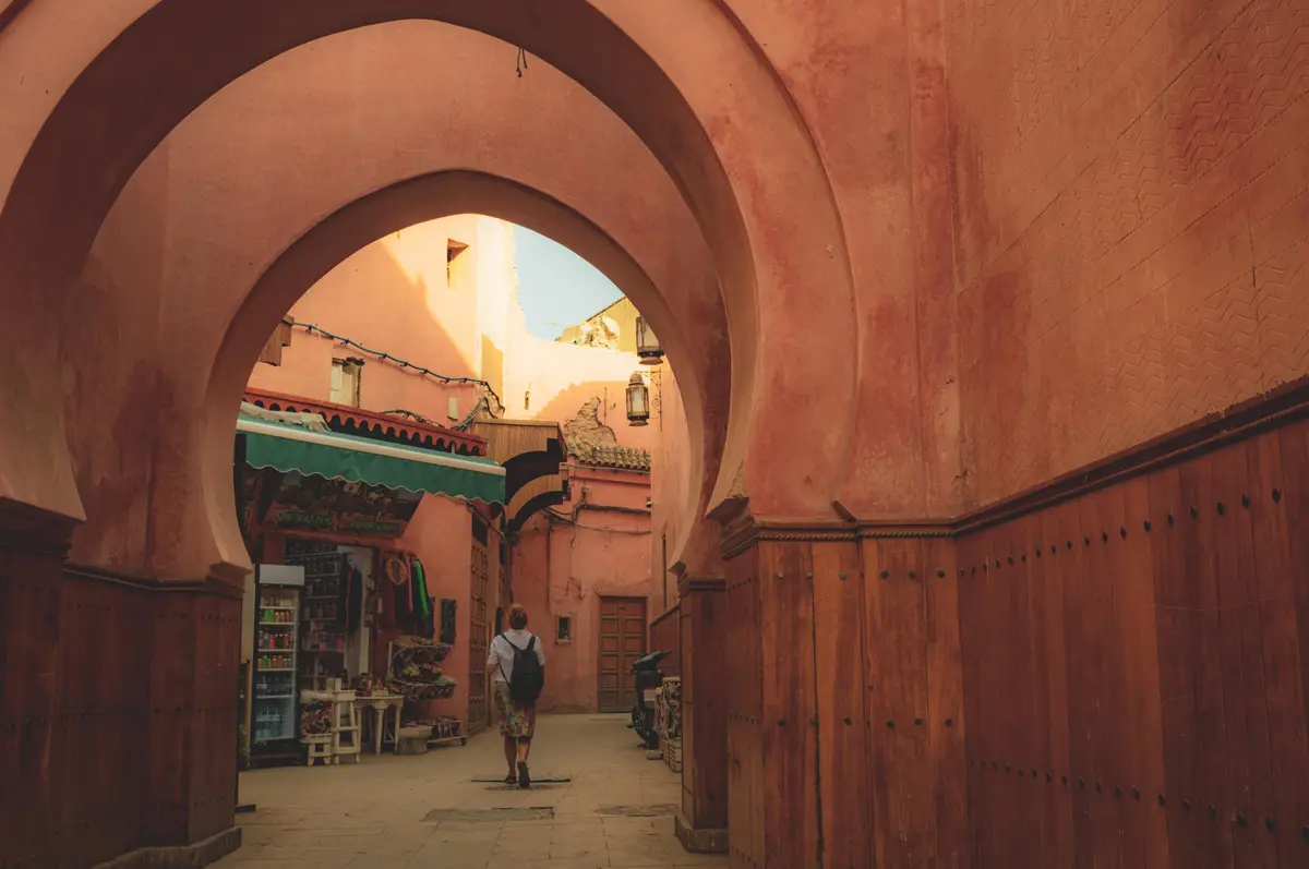 A person with a backpack walks away down a narrow alleyway framed by large, reddish-pink arched doorways. The alley is lined…