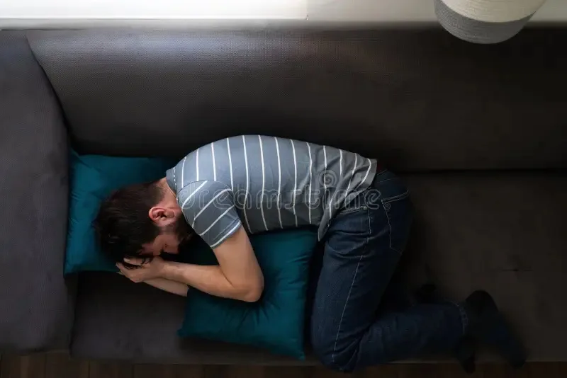 a man is seen from above, kneeling on a dark gray couch with his head buried in a teal pillow, wearing a gray striped…