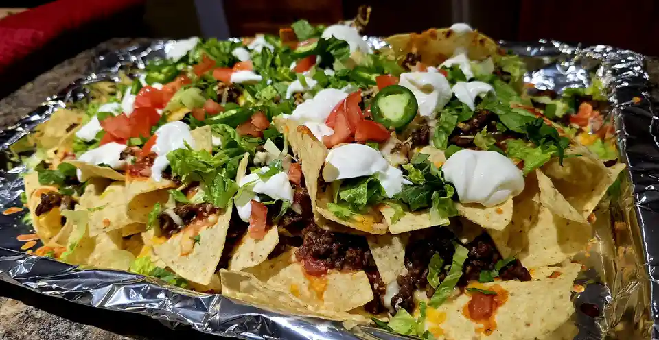A close-up photo shows a large tray of loaded nachos on aluminum foil, topped with melted cheese, ground meat, shredded…