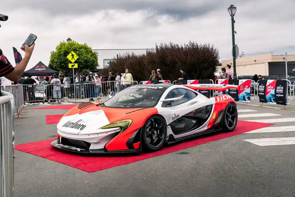 A red and white McLaren supercar with 'Marlboro' branding is parked on a red carpet at an outdoor car event, surrounded by…