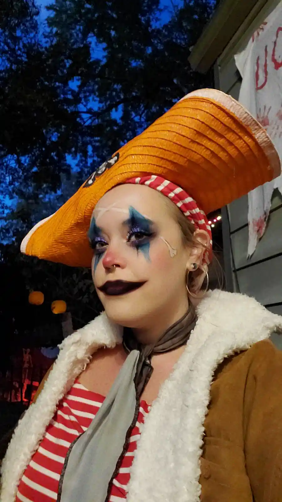 A person dressed as a pirate with clown makeup, wearing a large orange hat, a red and white striped shirt, and a brown…