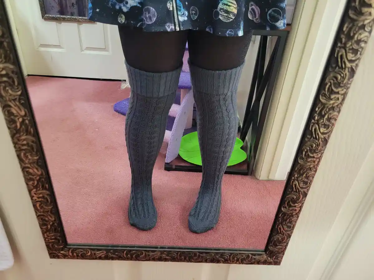 A mirror selfie shows a person wearing dark grey, cable-knit, knee-high socks over black tights, paired with a space-themed…