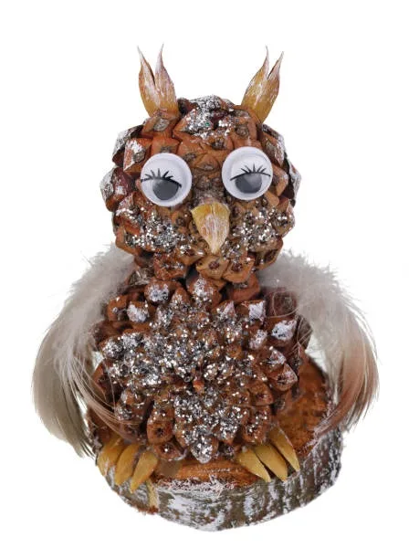 A decorative owl crafted from pinecones, glitter, and feathers, with large googly eyes and feathered wings, sits on a birch…