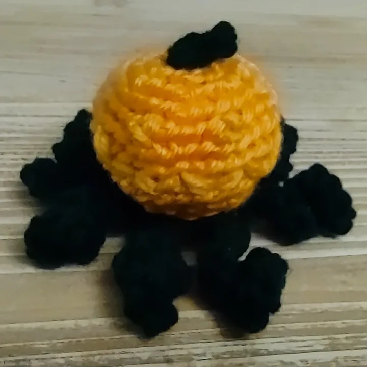 A handmade crocheted pumpkin made of orange yarn with a black yarn stem, but it has black yarn legs attached, giving it the…