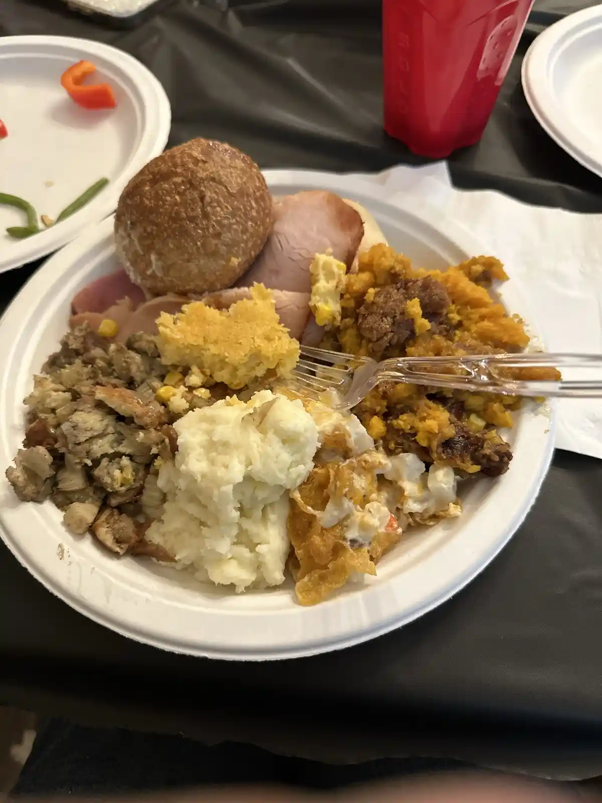 A close-up photo shows a white paper plate piled high with various Thanksgiving-style foods including ham, mashed potatoes…