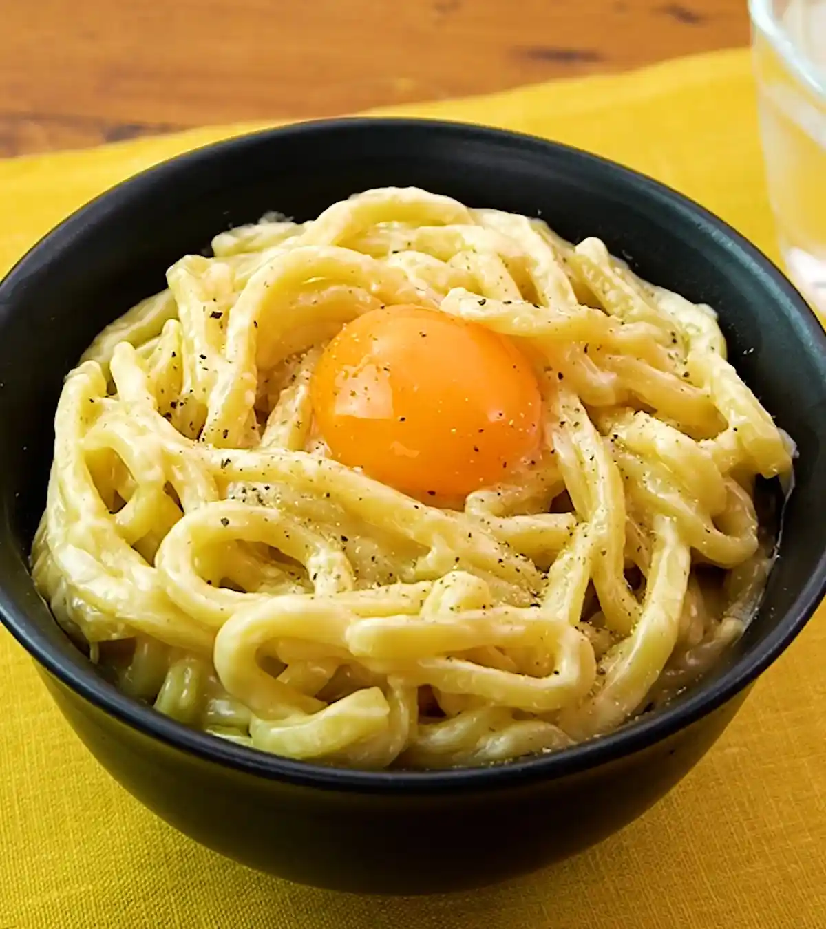 A close-up photo shows a black bowl filled with creamy pasta, topped with a single, intact raw egg yolk and sprinkled with…