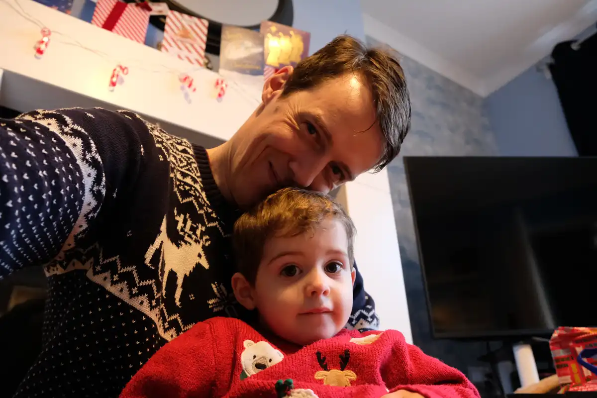 A man in a dark blue holiday sweater with white reindeer patterns leans in to take a selfie with a young boy in a red…