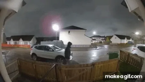 A security camera captures a person walking toward a white SUV at night, then suddenly crouching low to the ground as if…