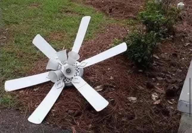 A white ceiling fan with five blades lies on its side in a grassy yard with mulch and a bush nearby. The overlaid text reads…