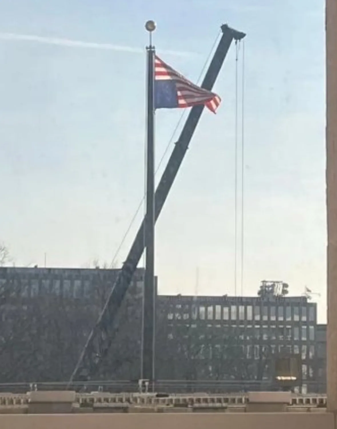 An american flag is visibly being lowered from a flagpole by the boom of a large construction crane, with city buildings in…