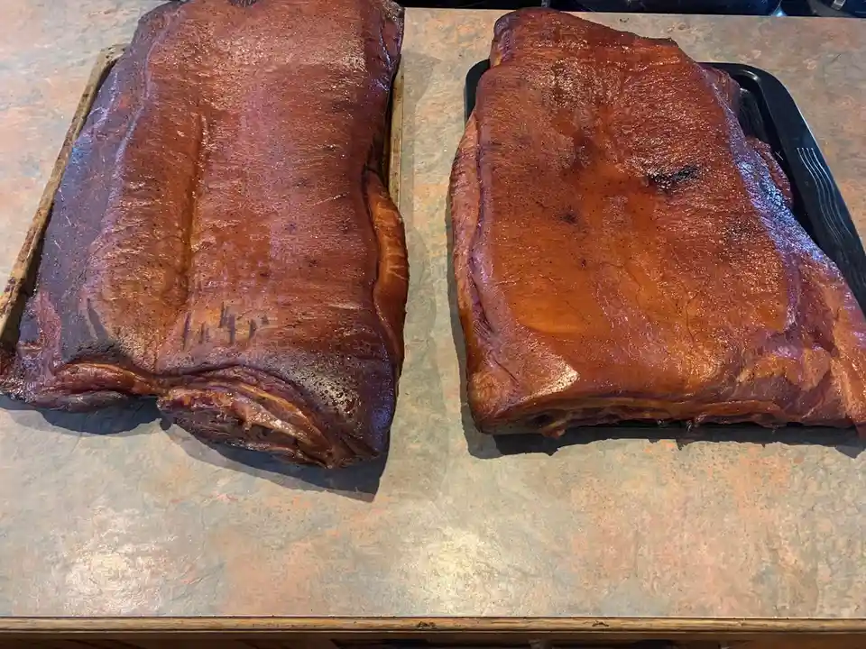 Two large, glistening, smoked pork ribs sit side-by-side on baking trays atop a speckled countertop. The left slab has…