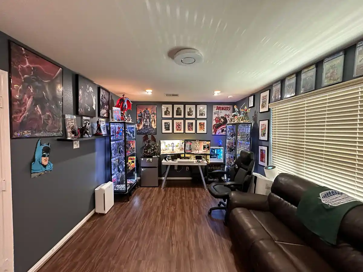 A wide shot of a meticulously decorated gaming and comic book room with dark walls, wooden floors, and a brown leather…
