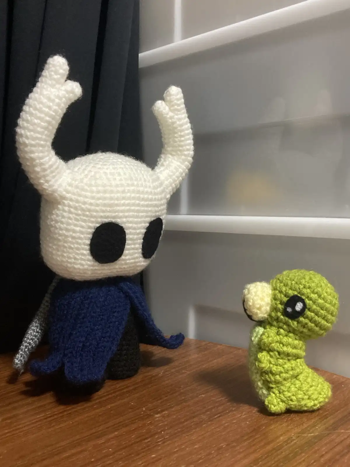 Two crocheted plush toys sit on a wooden surface: a larger white figure with horns and a blue cape, and a smaller green…