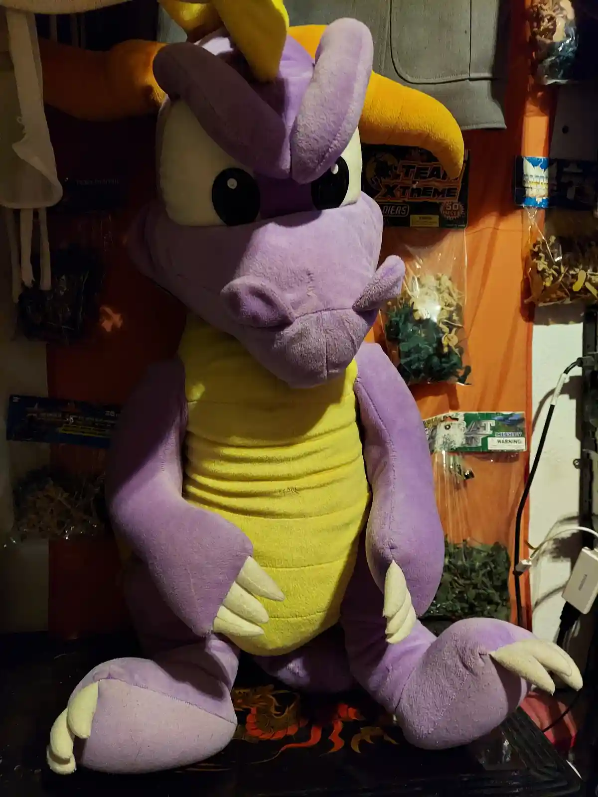 A large purple and yellow plush dragon toy, resembling Spyro the Dragon, sits upright on a surface with a slightly smug or…