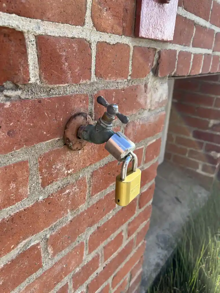 A close-up photo shows a metal outdoor faucet protruding from a red brick wall, with a yellow padlock securing its handle…