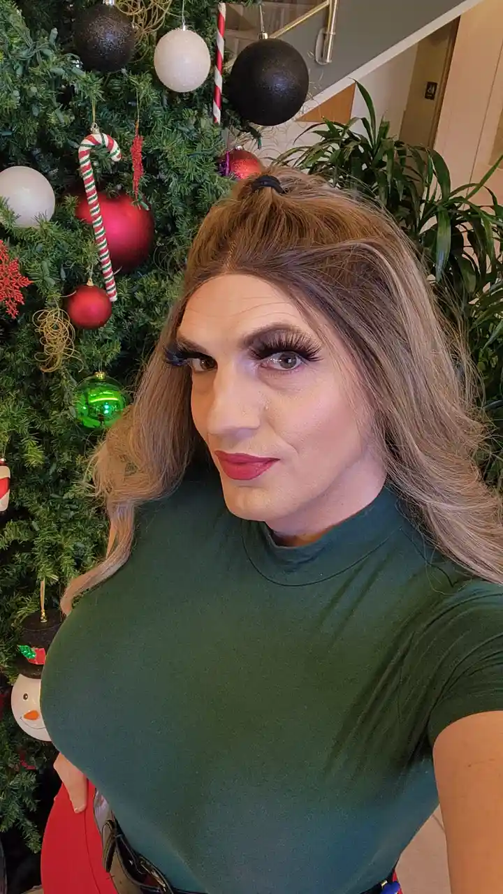 A person with long blonde hair, heavy makeup including thick false lashes and red lipstick, and wearing a green turtleneck…