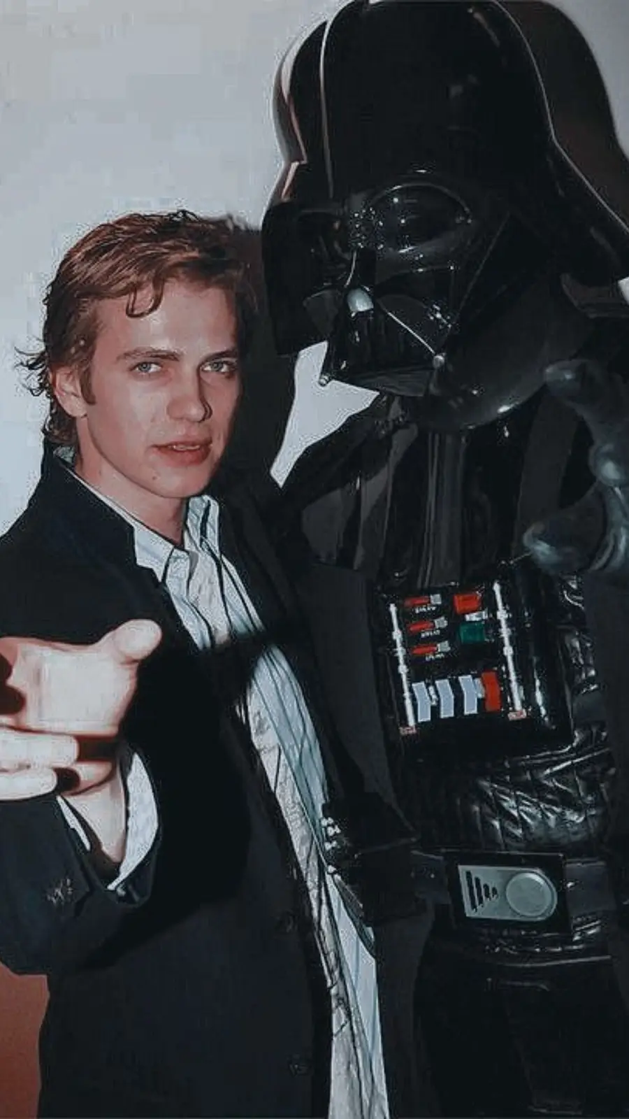 A man in a suit points forward while standing next to a full-body Darth Vader costume. The man has a neutral expression. The…