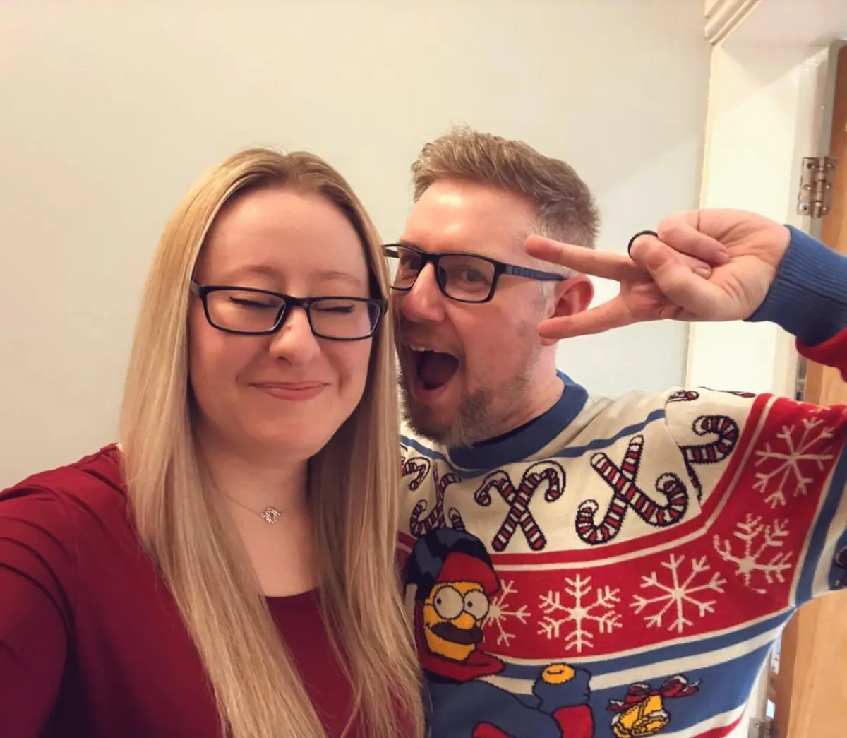 A selfie of a man and woman; the man wears a festive, overly decorated Christmas sweater featuring Homer Simpson and candy…