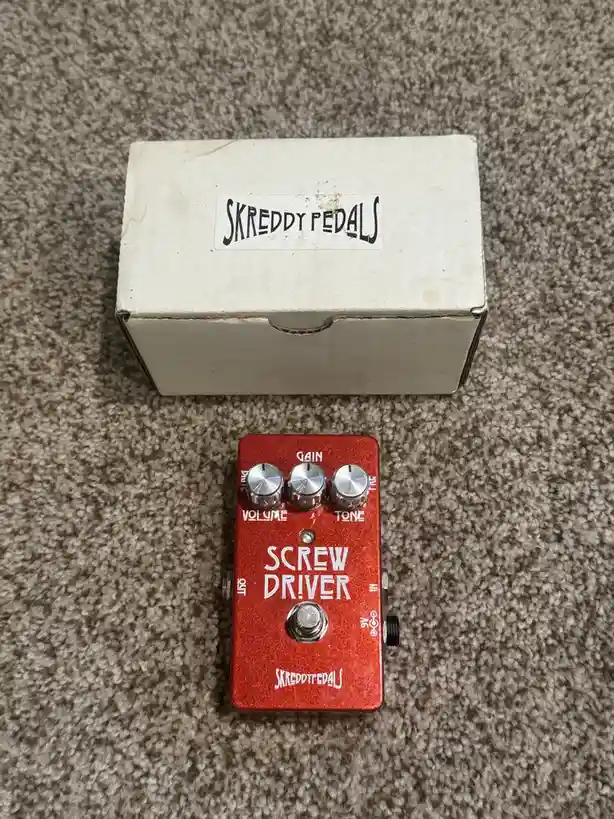 A red guitar effects pedal labeled 'SCREW DRIVER' by 'SKREDDY PEDALS' sits on carpet next to its white cardboard box, which…