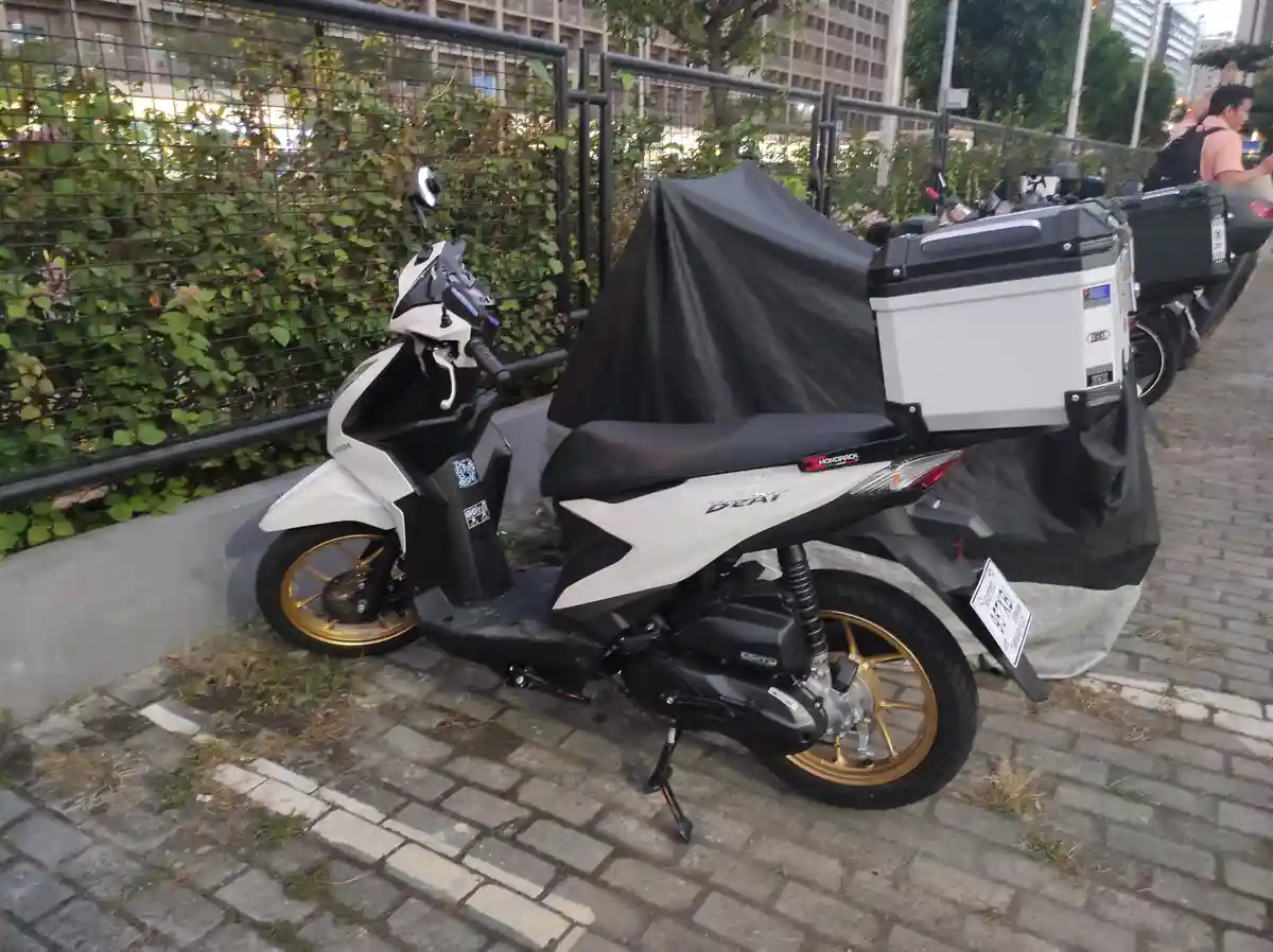 A white and black Honda Beat scooter with gold wheels is parked on a brick sidewalk, partially covered by a black tarp that…